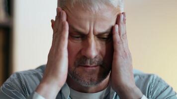 A middle-aged man holds his head in his hands, looking distressed and overwhelmed while seated at a desk in a warmly lit room. video