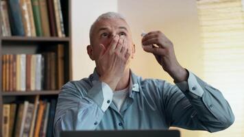 A man uses eye drops while seated at a desk, surrounded by shelves filled with books, ready for online class video