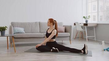 A woman dressed in activewear performs a stretching exercise on a yoga mat in a stylish living room. The bright space features a sofa, plants, and exercise equipment nearby. video