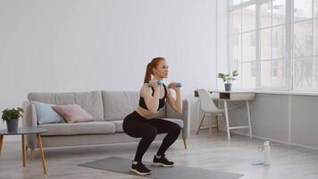 A woman performs strength training with dumbbells in a sunlit living space. Surrounded by modern decor, she focuses on her fitness routine while maintaining proper form and balance. video