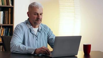 uma homem com cinzento cabelo é focado em dele computador portátil enquanto sentado às uma mesa, com caloroso iluminação e uma caneca próximo. video