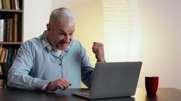 A man dressed in pajamas is focused on his laptop, seated at a table in a warmly lit room with bookshelves behind him. video