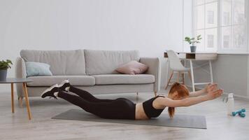 A woman practices floor exercises at home, extending her arms while lying on a yoga mat. The bright, spacious room features a couch and a table. video