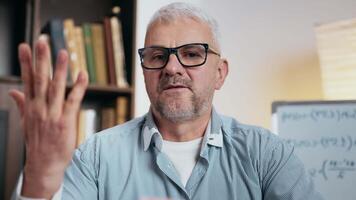 A man teaches math concepts online, gesturing with his hands while seated in a well-lit study surrounded by books. video