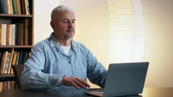 a Mais velho homem focos atentamente enquanto digitando em uma computador portátil às uma mesa, cercado de livros dentro uma esquentar, bem iluminado sala. video