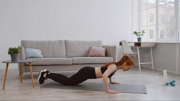 A fit young woman performs push-ups on a mat in her spacious living room. The area is well-lit with natural light, featuring a couch and a small workout space. video