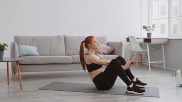 A young woman is exercising on a yoga mat in her living room, focusing on strengthening her core. The space features a cozy couch and plants, creating a serene workout atmosphere. video