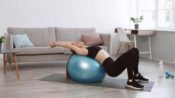 A woman performs a back stretch using a stability ball in a well-lit living room. The space is modern and inviting, with a couch and plants in the background. video