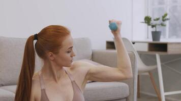 A woman with red hair is performing an arm exercise with a dumbbell in a cozy living room. Sunlight streams through the window, illuminating her focused expression. video