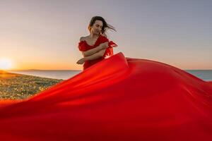 A woman in a red dress is standing on a grassy hillside. The sun is setting in the background, casting a warm glow over the scene. The woman is enjoying the beautiful view and the peaceful atmosphere. photo