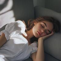 A young woman in a white outfit sleeps on a grey sofa, with soft light casting shadows, creating a calm atmosphere. photo