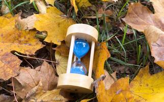 Hourglass with autumn leaves on bench in park, closeup photo
