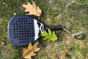 Top view of a black padel racket on a green court grass turf behind the net outdoors. Paddle is a racquet game. Professional sport concept with space for text photo