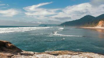 Coastal beach with waves and distant rocky cliffs. A beautiful coastal scene with waves gently rolling onto the beach, bordered by forested cliffs in the distance. video
