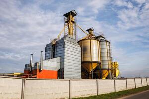 large grain elevator with multiple cylindrical metal silos, structural elements, and a complex system of pipes. It stands against a blue sky, with a field in the foreground after harvest photo
