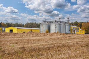 large grain elevator with multiple cylindrical metal silos, structural elements, and a complex system of pipes. It stands against a blue sky, with a field in the foreground after harvest photo