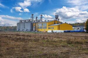 large grain elevator with multiple cylindrical metal silos, structural elements, and a complex system of pipes. It stands against a blue sky, with a field in the foreground after harvest photo