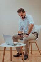 A confident businessman sitting and using laptop and smartphone with a determined expression, while a beige background enhances the professional atmosphere, showcasing his productivity and expertise. photo