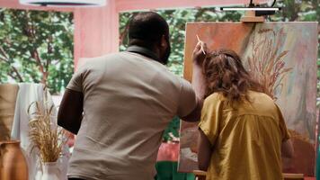 Talented artist and her apprentice applying watercolor paint on canvas, working together to create beautiful nature inspired masterpiece in atelier. Two people painting colorful scene. Camera A. video
