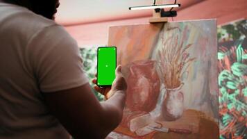 Young artist looking at isolated chroma key display in his painting atelier, using device next to canvas with colorful masterpiece. Painter creating rustic artwork for gallery exhibition. Camera B. video