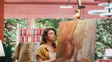 Female painter using watercolor paint and mixing colors on palette for her artistic creation in fine art workshop. Woman painting rustic elements on canvas, creative process in atelier. Camera A. video