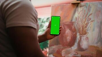 African american artist holding device with green screen mockup in art studio, working on a new painting with a certain technique. Painter using watercolor and acrylic dye for his creation. Camera B. video