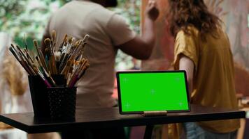 Diverse artists painting new artwork next to isolated mockup screen, collaborating on masterpiece for fine art gallery exhibition. Painter and apprentice using watercolor paints on canvas. Camera A. video