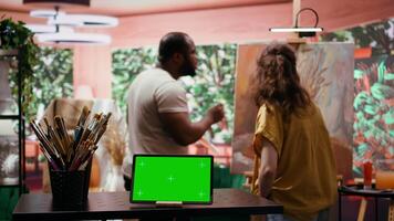 Two artists painting on a canvas next to green screen on tablet display, working together on their new masterpiece for fine art gallery exhibition. Diverse team of painters in art studio. Camera B. video