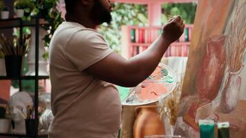 Focused man painting a new rustic artwork on canvas with brushstrokes patterns, mixing watercolors and applying to create a masterpiece. Young male artist working on his creation. Camera A. video