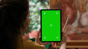 Artist using mockup copy space display in her painting workshop, looking at screen next to her rustic colorful masterpiece. Woman examining her finished artwork with watercolor paint. Camera A. video