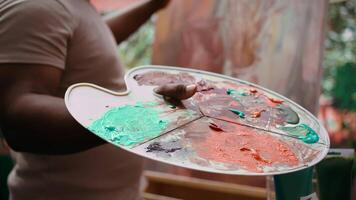 In an art studio, painter mixes multiple watercolor paints on a palette to create artwork on canvas. Skilled man painting with tools and showing imagination, artistic creation. Close up. Camera B. video