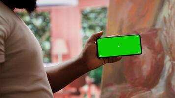 Young artist looking at isolated chroma key display in painting atelier, using device next to canvas with colorful masterpiece. Painter creating rustic artwork for the gallery exhibition. Camera A. video