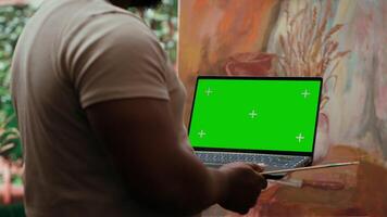 African american artist holding device with green screen mockup in art studio, working on a new painting with a certain technique. Painter using watercolor and acrylic dye for his creation. Camera A. video