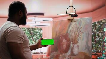 African american painter using a gadget with copy space screen in workshop, working on his new nature inspired masterpiece on canvas. Using watercolor paint and creativity in atelier. Camera B. video