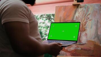 African american artist holding device with green screen mockup in art studio, working on a new painting with a certain technique. Painter using watercolor and acrylic dye for his creation. Camera B. video