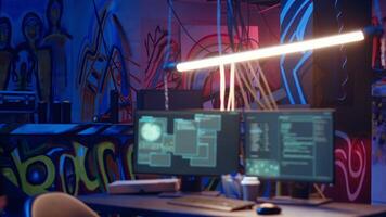 Hooded hacker arrives in neon lit underground shelter filled with graffiti artwork. Cybercriminal enters colorful painted basement, starting work on script that can crash businesses servers, camera B video