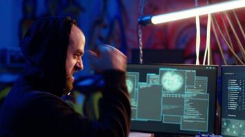 Hooded hacker running code in graffiti painted basement, looking to find exploits for system vulnerabilities to be used against system owner for ransom, camera B handheld camera shot video