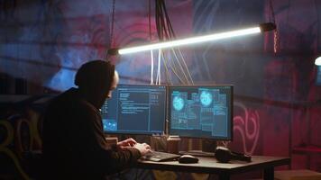 Hacker arrives in messy base of operations with neon lights and graffiti drawings sprayed on walls. Cybercriminal using ghetto hideout as HQ to commit illegal activities using computer, camera B video