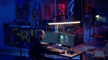 Hacker in underground warehouse writing lines of code on computer screen to make malware that get past security systems. Man in hidden HQ building scripts that can hack devices, dolly in shot video
