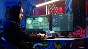 Hooded hacker running code in graffiti painted basement, looking to find exploits for system vulnerabilities to be used against system owner for ransom, camera A handheld camera shot video