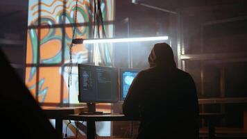 Hacker using software interface on computer screen, running programming code in warehouse with graffiti sprayed on walls. Evil developer in underground base building malware, handheld camera shot video