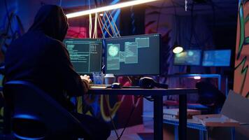 Hooded man in underground hideaway listening and breaking into computer systems to access valuable data. Evil hacker wearing headphones bypassing firewalls and login process, panning shot video