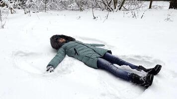 Happy woman lying on snowy lawn at forest and making angel. Young girl playing in snow and enjoying wintertime. Cheerful lady having fun outdoor. Concept of a winter vacation. Close up Slow mo video