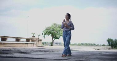 Handheld low angle view shot, A cheerful Asian woman enjoys drink iced coffee and takes a leisurely stroll along a concrete walkway in a park. Weekend relaxation time video