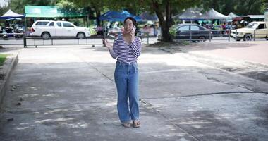 Handheld shot, A cheerful Asian woman takes a leisurely stroll along a concrete walkway while talking with friend on smartphone in a park. Weekend relaxation time video