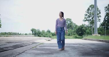 Handheld low angle view shot, A cheerful Asian woman enjoys the peaceful atmosphere and takes a leisurely stroll along a concrete walkway in a park. Weekend relaxation time video