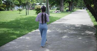 Handheld back view shot, A cheerful Asian woman enjoys the peaceful atmosphere and takes a leisurely stroll along a concrete walkway in a park. Weekend relaxation time video