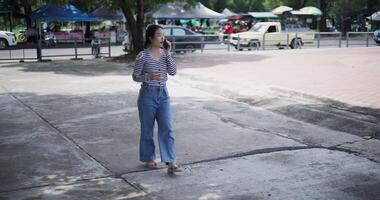 Handheld shot, A cheerful Asian woman takes a leisurely stroll along a concrete walkway while talking with friend on smartphone in a park. Weekend relaxation time video
