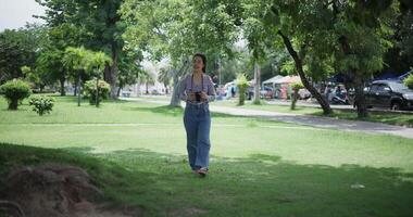 Handheld shot, Cheerful woman is walking in a park on a sunny day, surrounded by lush green trees, and enjoys taking pictures of the nature with her camera. A relaxing weekend moment. video