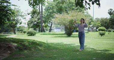 Handheld shot, Happy young woman is talking with her friend via call on smartphone while taking a leisurely stroll, surrounded by lush green trees in a park. Enjoy holiday activities video